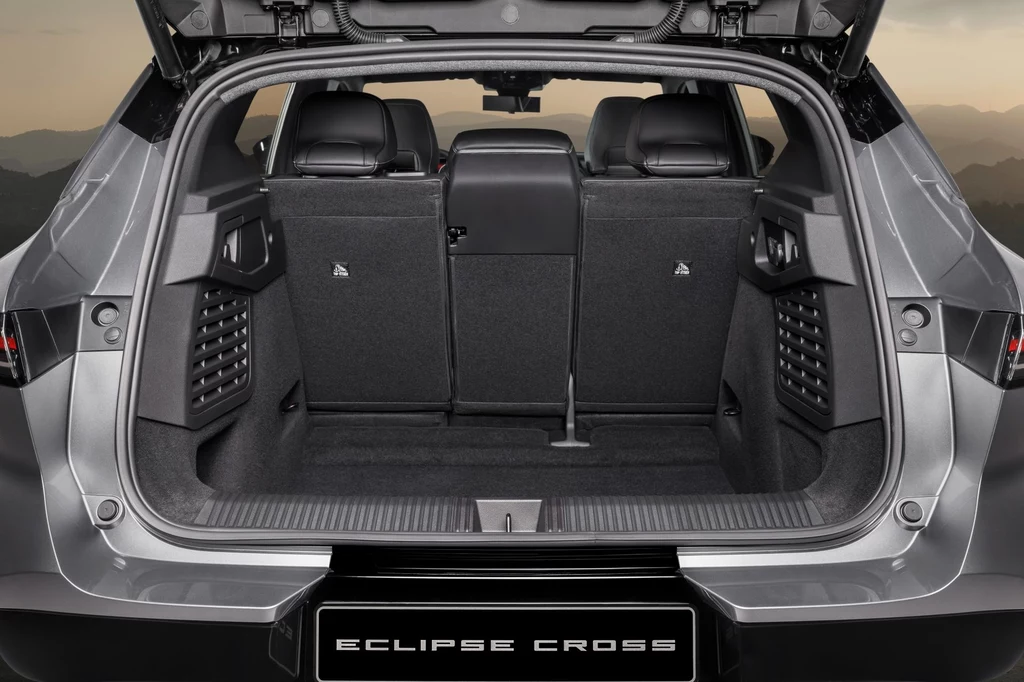 Bagażnik Mitsubishi Eclipse Cross pomieści 478 litrów. Po złożeniu tylnych siedzeń ta przestrzeń rośnie do 1 670 litrów. Bagażnik samochodu SUV z otwartą klapą, widoczne trzy tylne siedzenia oraz wykończenie wnętrza utrzymane w ciemnych odcieniach, po bokach wbudowane schowki i panele z tworzywa, logo Eclipse Cross na progu bagażnika.