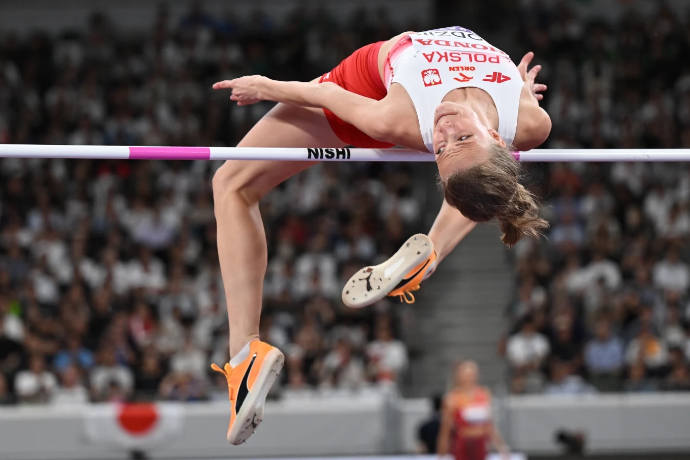 Lekkoatletka w stroju reprezentacji Polski wykonuje skok wzwyż, przekraczając poprzeczkę podczas zawodów na stadionie w obecności licznej publiczności.