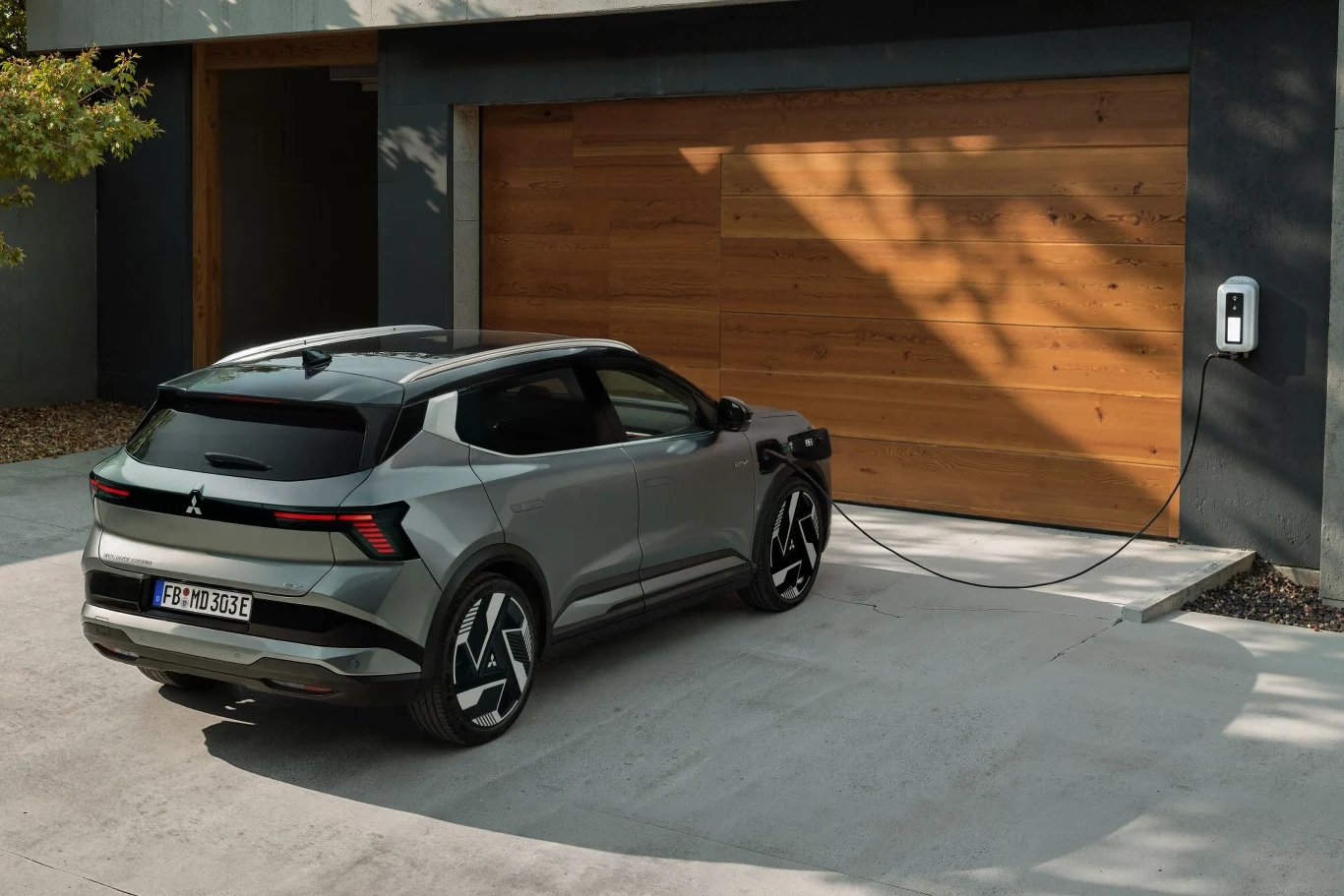 Nowoczesny srebrny samochód elektryczny typu SUV podczas ładowania na podjeździe przed drewnianą bramą garażową, samochód podłączony do domowej stacji ładowania zamontowanej na ścianie budynku.