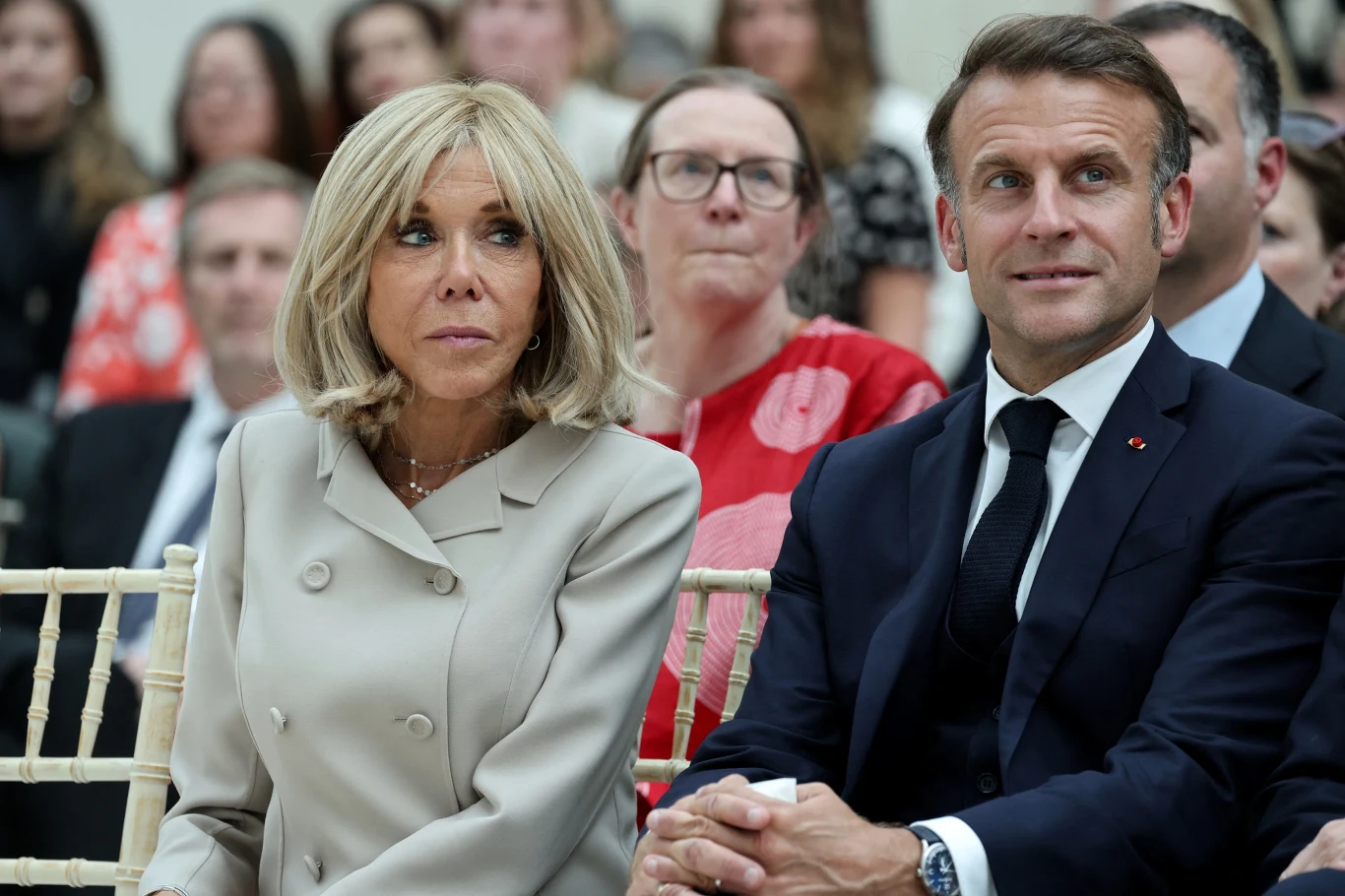 Brigitte i Emmanuel Macron mają dowieść przed sądem, że pierwsza dama jest kobietą Para elegancko ubranych osób siedzi na krzesłach wśród publiczności, skupiając uwagę na czymś poza kadrem; w tle widoczne są inne osoby obserwujące wydarzenie.