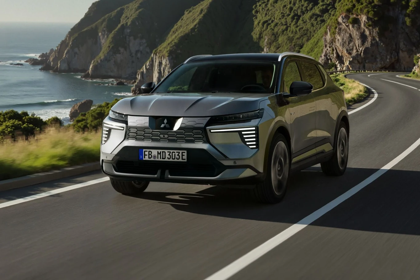 Nowoczesny, srebrny SUV Mitsubishi poruszający się po krętej, asfaltowej drodze z widokiem na morze, klify i bujną roślinność w tle.