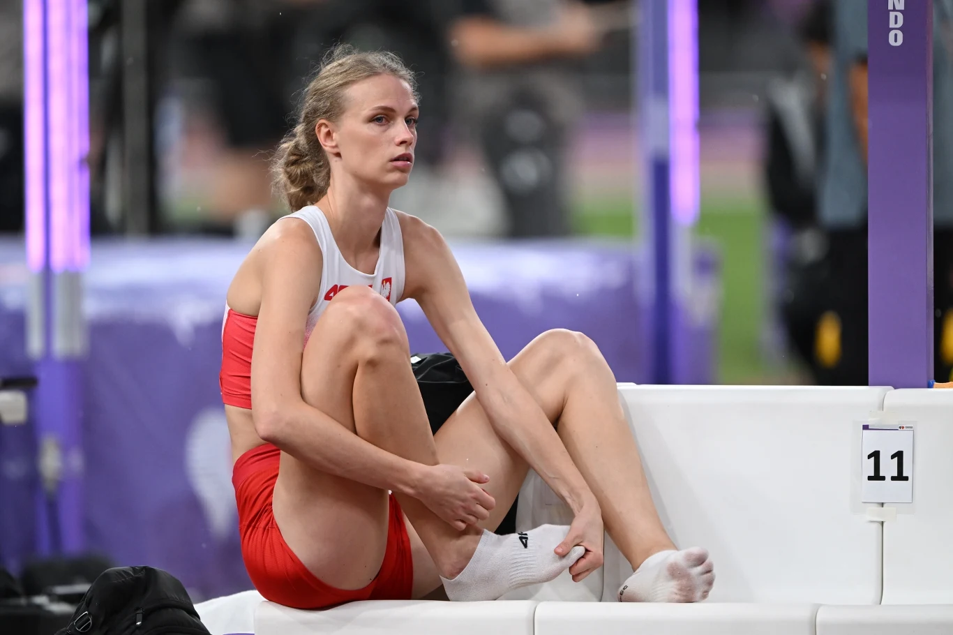 Sportsmenka w stroju sportowym czerwono-białym siedzi na białej ławce na stadionie, wygląda na zmęczoną lub zamyśloną, z opatrzoną nogą, w tle rozmyte elementy hali sportowej.