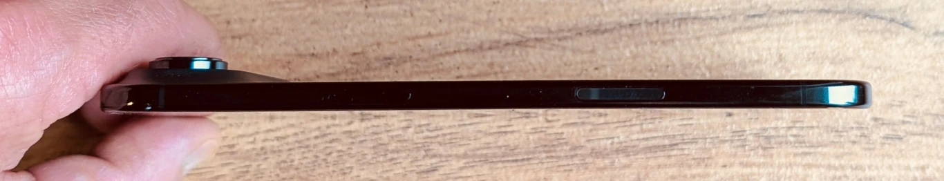 Czarny, bardzo cienki smartfon trzymany w dłoni, widok z boku na płaski profil urządzenia oraz wystający obiektyw aparatu na tle drewnianej powierzchni.