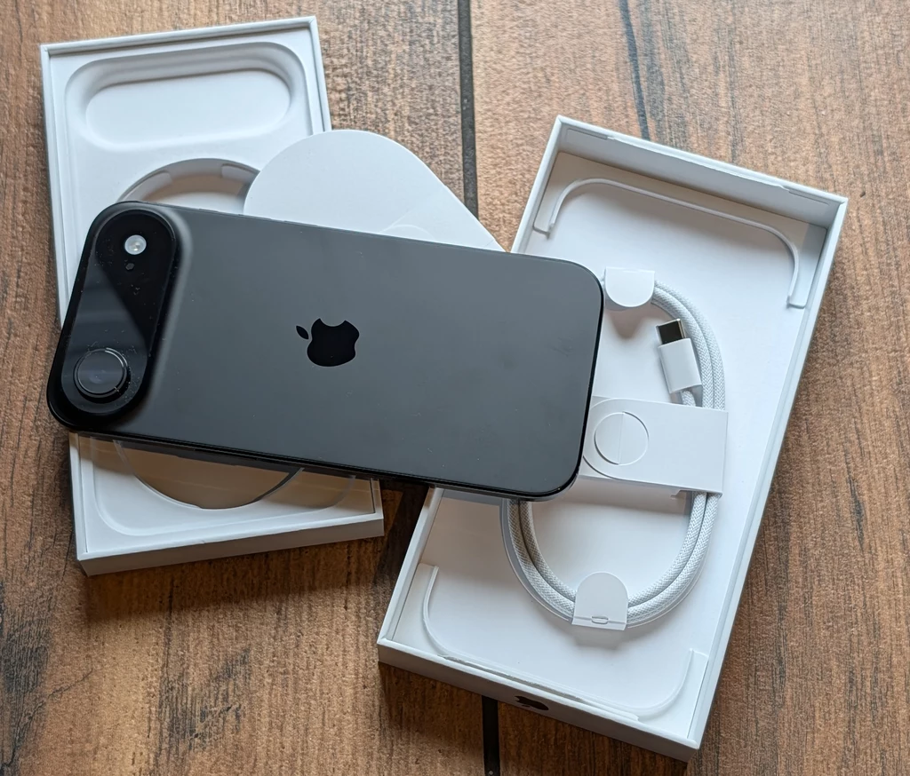 Czarny smartfon marki Apple położony na otwartym opakowaniu z widocznym kablem do ładowania USB-C umieszczonym w pudełku, całość znajduje się na drewnianej powierzchni.