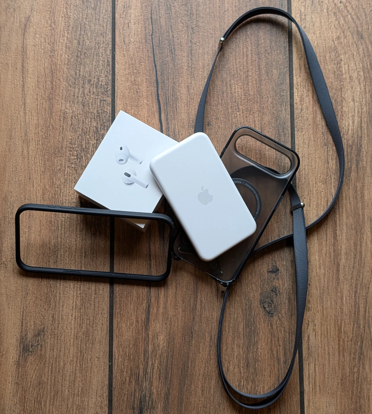 Białe bezprzewodowe słuchawki AirPods w oryginalnym opakowaniu, powerbank MagSafe Apple, dwa czarne etui na telefon i czarny pasek leżą na drewnianej podłodze.