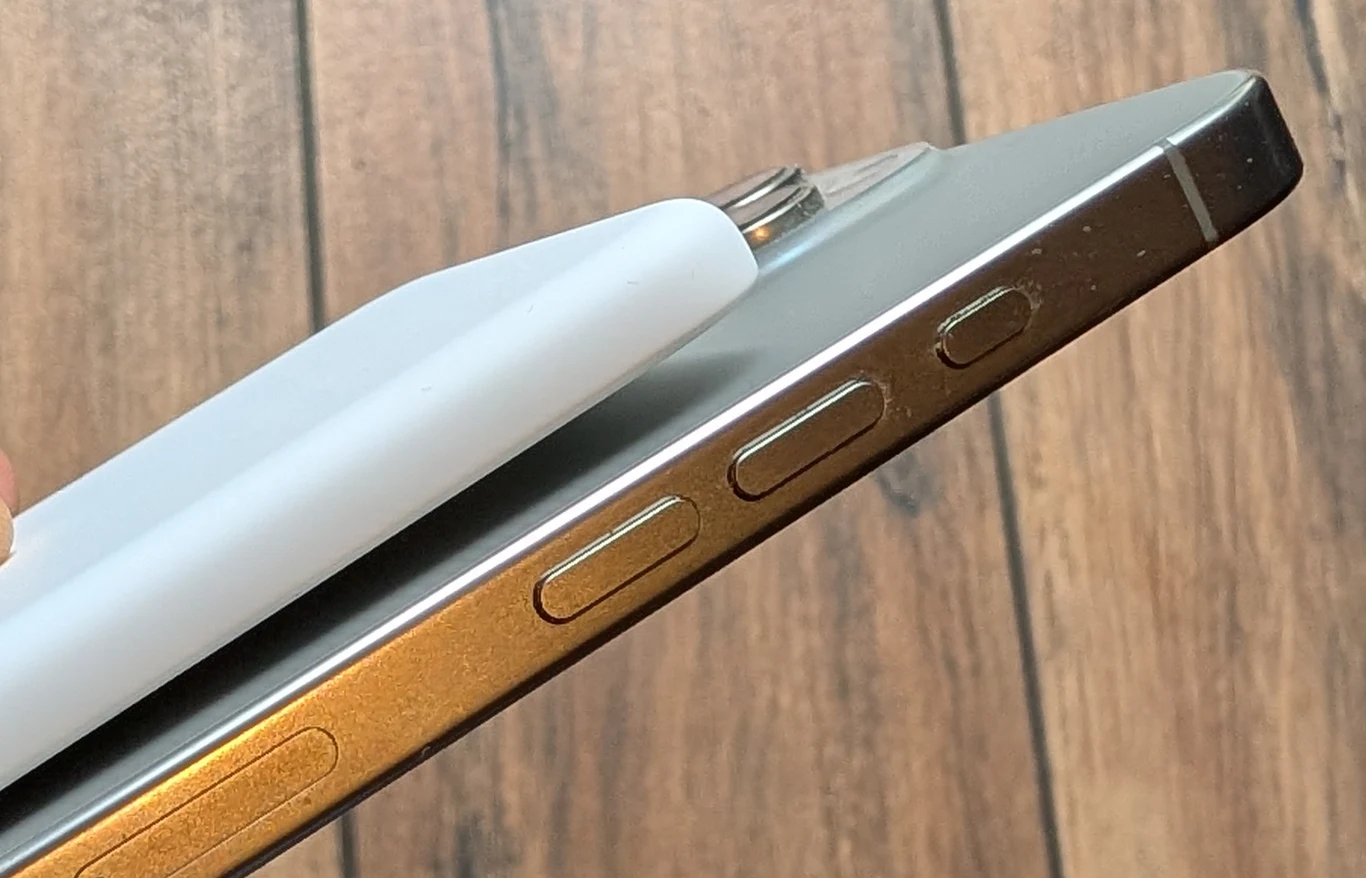 Zbliżenie na bok nowoczesnego smartfona oraz stylus magnetycznie przyczepiony do powierzchni urządzenia, na tle drewnianej podłogi.