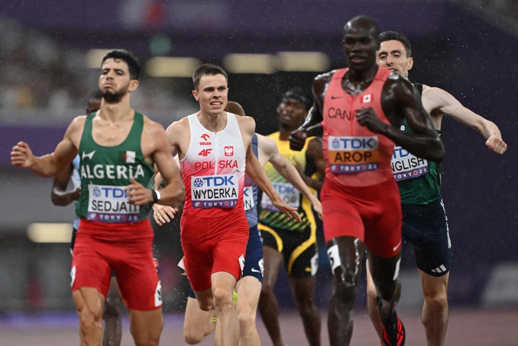 Maciej Wyderka za wielkimi mistrzami dystansu 800 metrów Grupa lekkoatletów biegnących na zawodach, wyraźnie widoczni zawodnicy z Algierii, Polski, Kanady oraz Wielkiej Brytanii, ubrani w narodowe stroje startowe, dynamiczna scena na stadionie lekkoatletycznym.