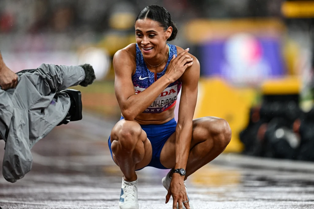 Sydney McLaughlin-Levrone po swoim zwycięstwie w finale mistrzostw świata Sportowiec w stroju lekkoatletycznym kuca na wilgotnej bieżni z szerokim uśmiechem na twarzy, z lewej strony widoczny kamerzysta filmujący wydarzenie.