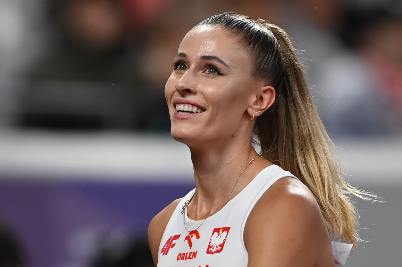 Uśmiechnięta lekkoatletka w biało-czerwonym stroju sportowym z widocznym godłem Polski i napisem Orlen na piersi, w tle rozmyte trybuny stadionu.
