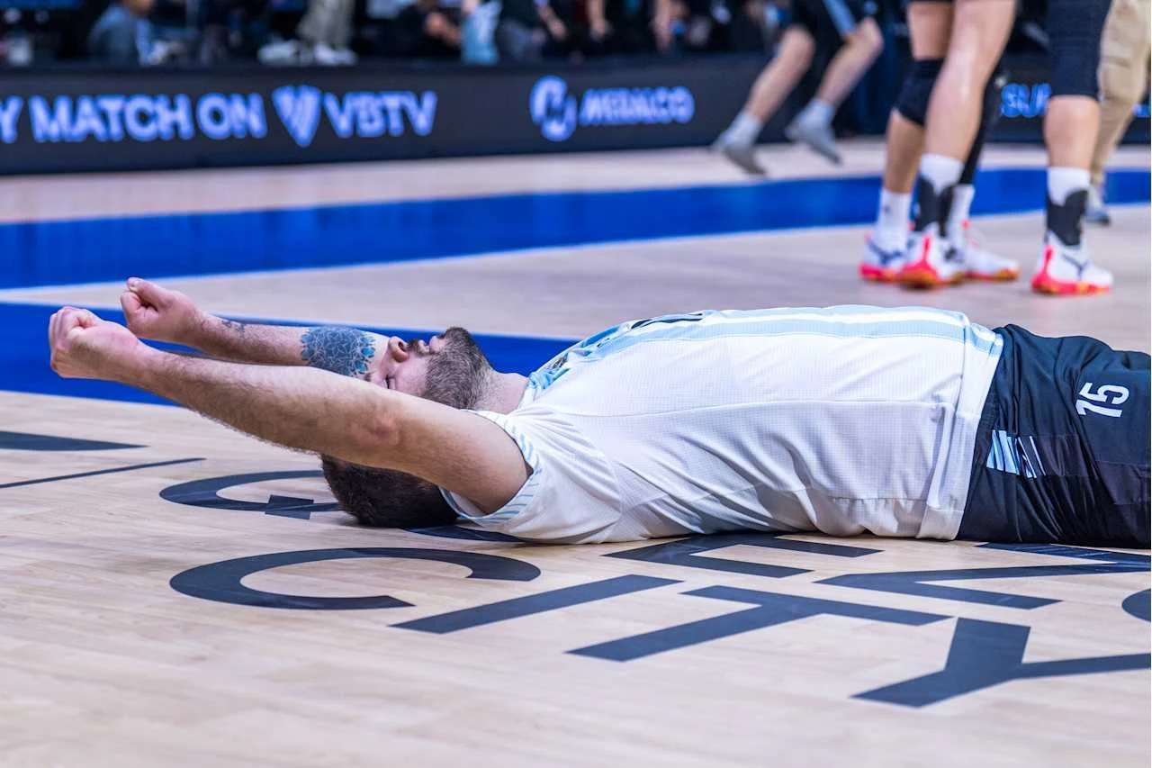 Siatkarz w białej koszulce leży na parkiecie z wyciągniętymi w górę rękami, prawdopodobnie okazując radość po zdobyciu punktu podczas meczu siatkówki, na tle wyraźnych oznaczeń boiska i innych graczy w tle.