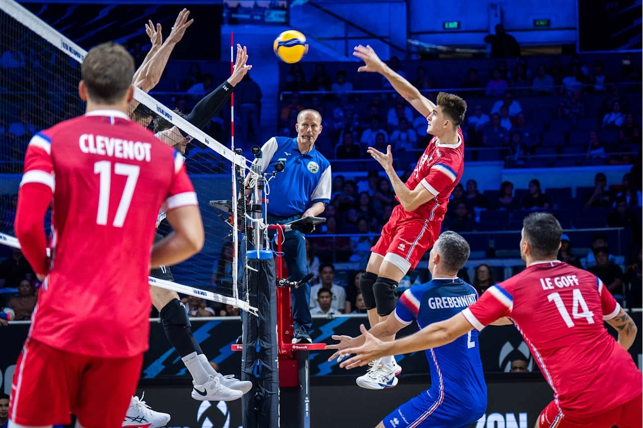 Siatkarze rywalizujący przy siatce podczas dynamicznej akcji meczu, z sędzią obserwującym rozgrywkę i widownią w tle, sportowcy w czerwonych i niebieskich strojach walczą o piłkę.