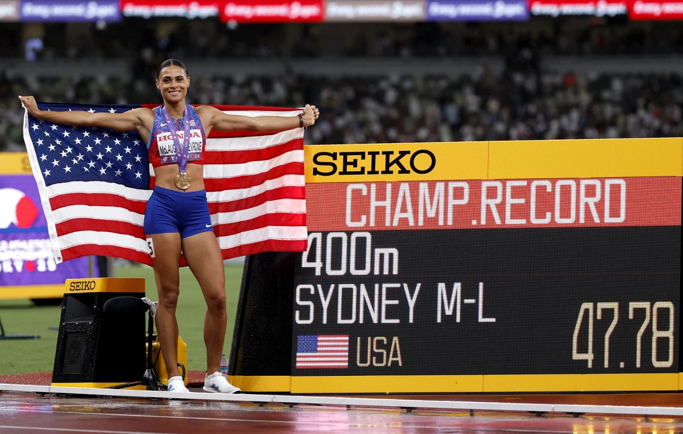 Lekkoatletka w sportowym stroju trzyma dużą flagę Stanów Zjednoczonych stojąc przy tablicy wyników z napisem 'CHAMP RECORD 400m SYDNEY M-L 47.78 USA', medal zawieszony na szyi, stadion w tle.