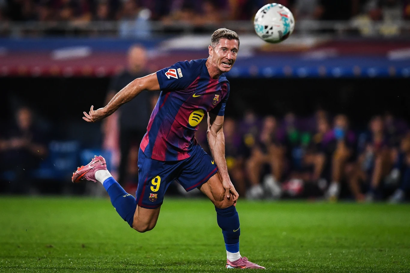 Robert Lewandowski Piłkarz w stroju FC Barcelony wykonuje dynamiczny ruch na boisku z piłką w powietrzu, intensywna akcja podczas meczu, w tle rozmazani kibice i ławka rezerwowych.