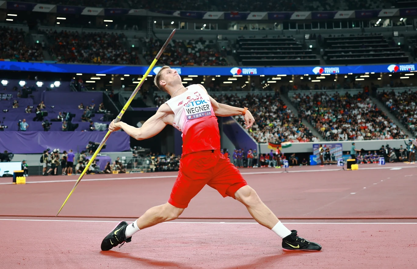 Sportowiec w białoczerwonym stroju wykonuje rzut oszczepem na stadionie lekkoatletycznym przy licznej widowni, skupiony i w napiętej, dynamicznej pozycji.