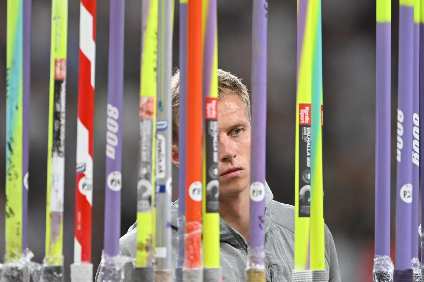 Zawodnik wpatruje się w stojak z kolorowymi tyczkami do skoku o tyczce, dominują pionowe linie sprzętu sportowego, tło niewyraźne, skupienie na twarzy sportowca i sprzęcie.