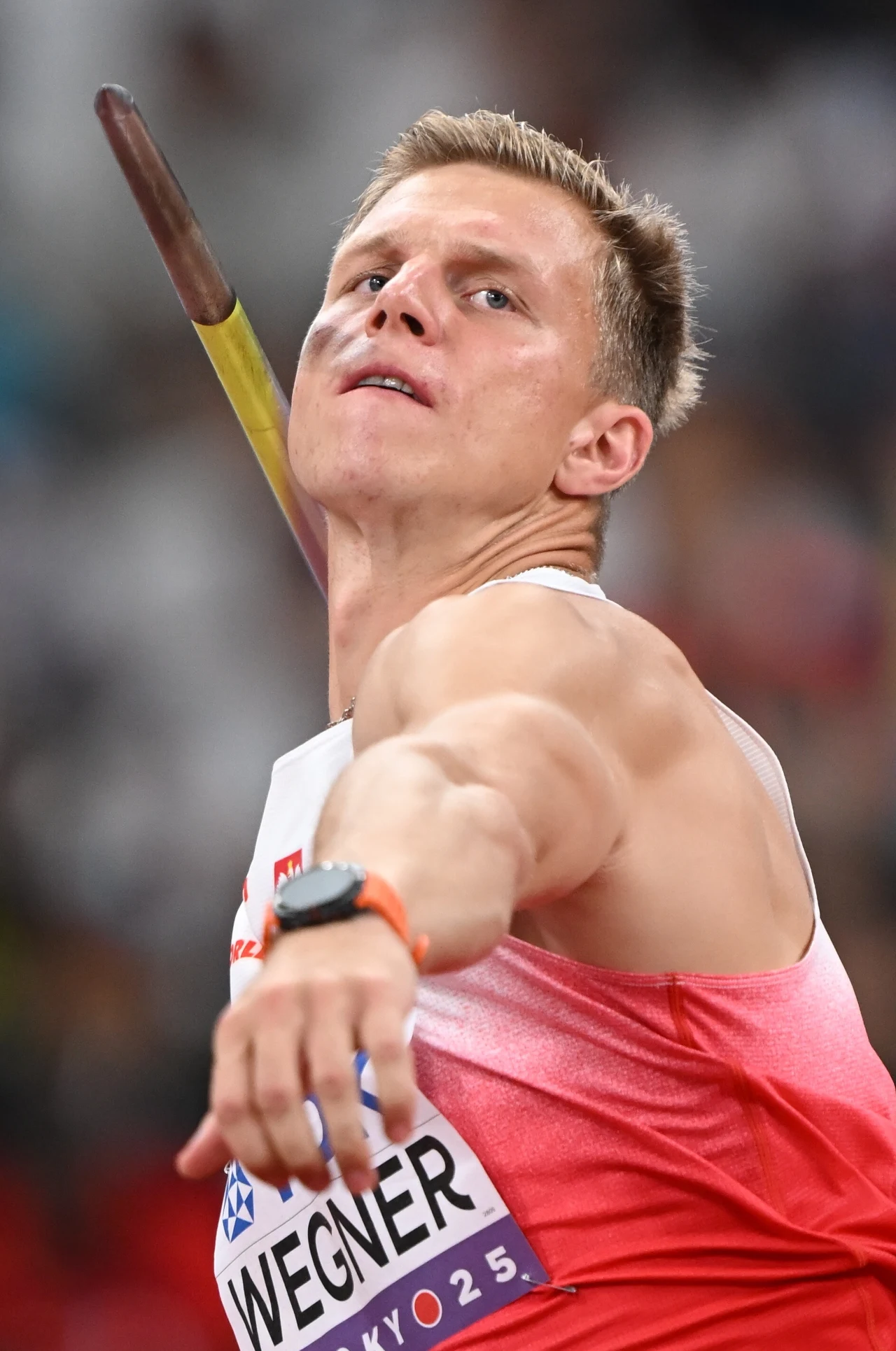 Sportowiec w dynamicznej pozie podczas rzutu oszczepem, skupiony wzrok, umięśnione ramię w trakcie wyrzutu, strój sportowy w biało-czerwonych barwach oraz numer startowy z nazwiskiem i oznaczeniem Tokio 2020.