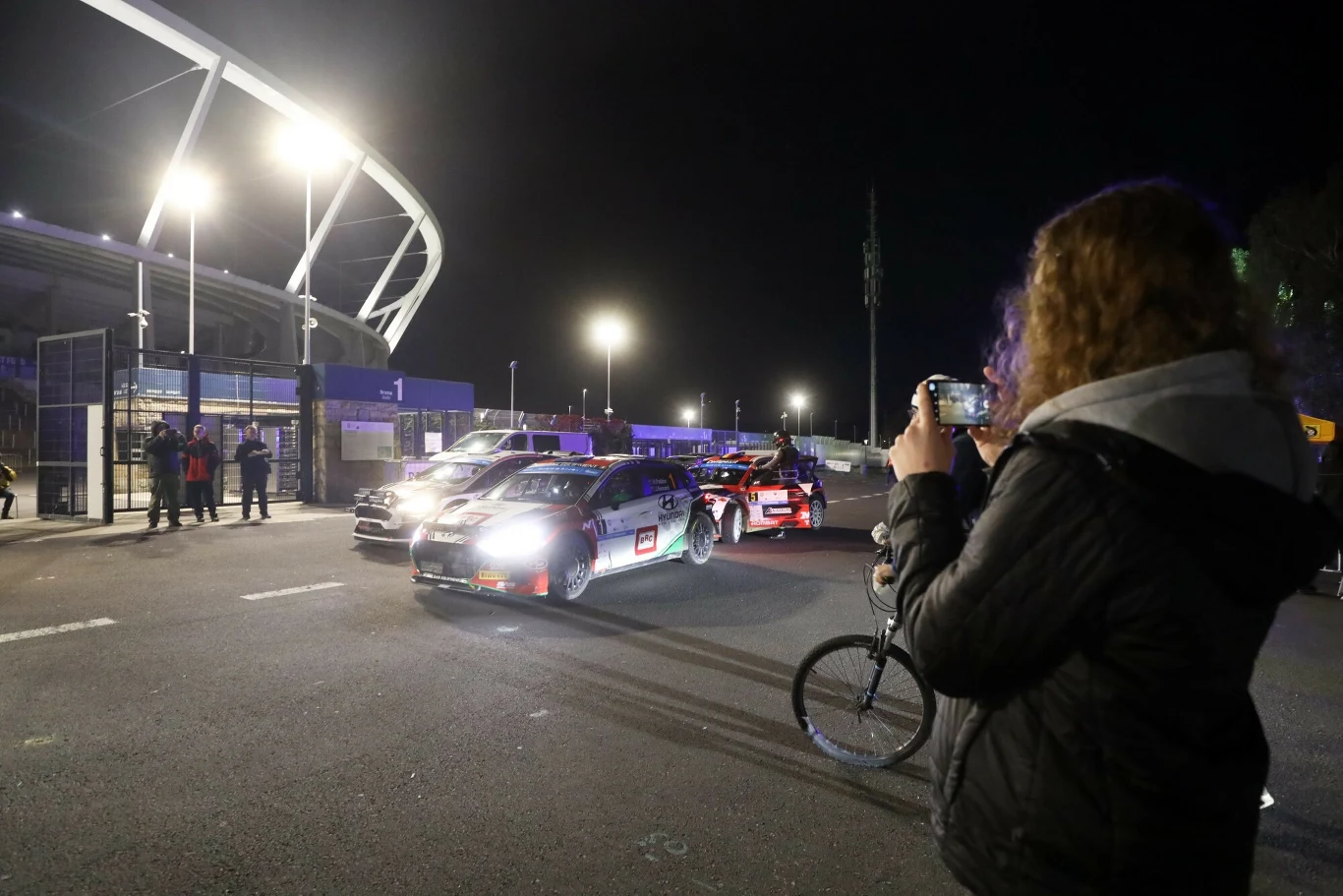 Rajd Śląska 2025. Organizatorzy proszą o rozwagę Rajdowe samochody ustawione na ulicy nocą podświetlone reflektorami, w tle stadion, z przodu kobieta fotografująca scenę telefonem, obok niej rower.
