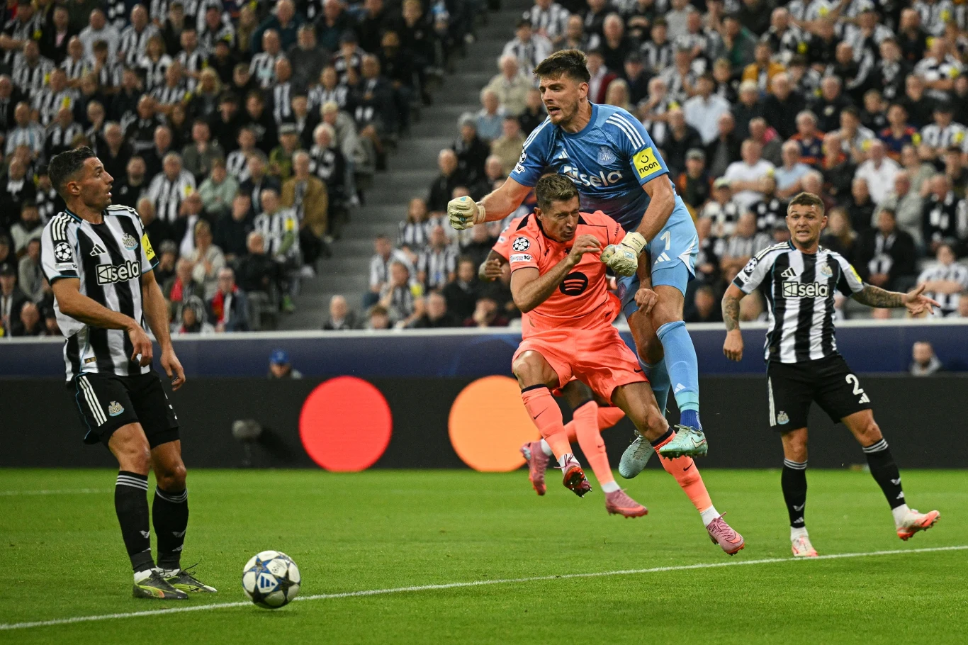 bramkarz w niebieskim stroju wyskakuje nad innym piłkarzem ubranym na różowo, starając się przechwycić piłkę, wokół znajdują się zawodnicy w czarno-białych strojach, a w tle widać trybuny pełne kibiców