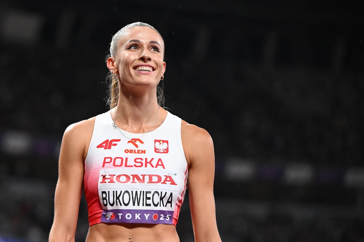 Młoda lekkoatletka w stroju reprezentacji Polski z nazwiskiem Bukowiecka na koszulce, stojąca na stadionie, uśmiechnięta, z widocznym logo sponsora oraz oznaczeniem igrzysk olimpijskich.