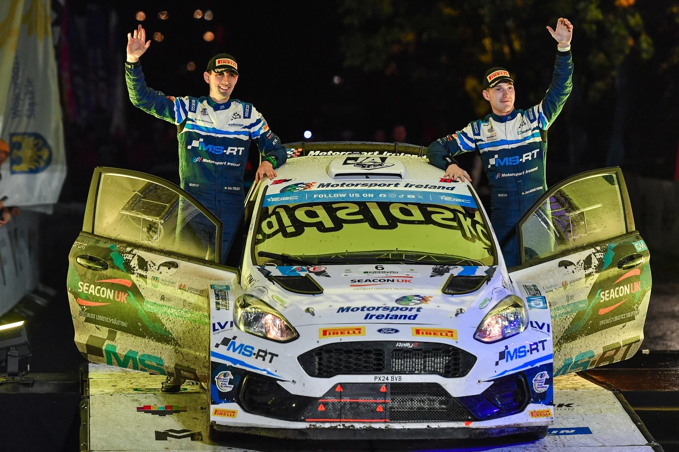 Rajd Śląska rozpocznie się w centrum Katowic Dwóch rajdowych kierowców w kombinezonach i z podniesionymi rękami stoi po obu stronach sportowego samochodu Ford Fiesta, którego drzwi są szeroko otwarte, celebrując sukces na mecie nocnego rajdu.