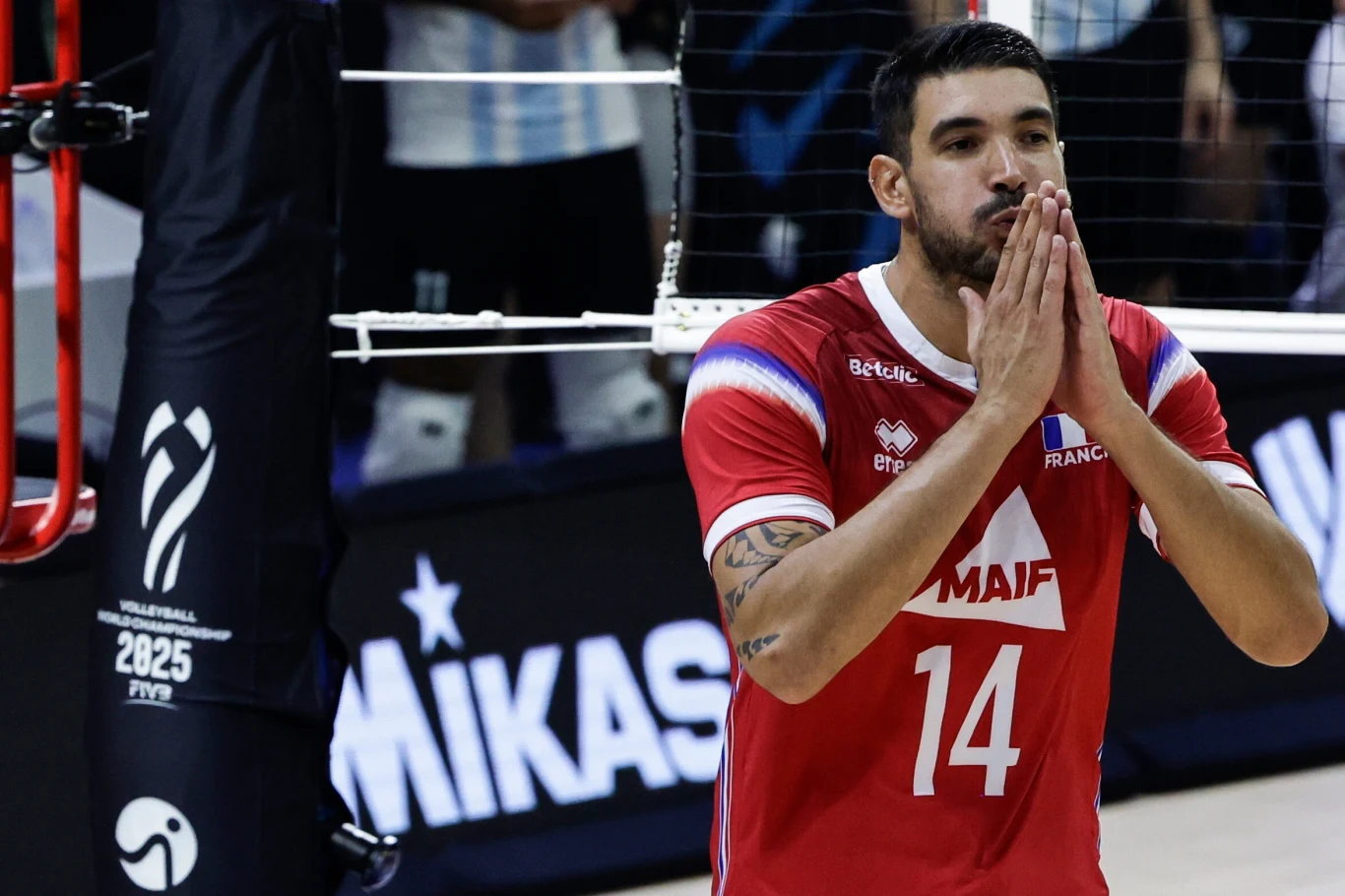 Siatkarz w czerwonym stroju z numerem 14 i logotypami sponsorów wykonuje gest złożonych dłoni na tle siatki i słupa z logo turnieju, w tle rozmazane elementy hali sportowej.