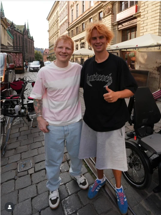 Dwóch uśmiechniętych młodych mężczyzn stoi razem na brukowanej ulicy miasta, obaj ubrani na luzie, jeden w jasnej, drugi w ciemnej koszulce. W tle widać zabudowania miejskie i rowery.