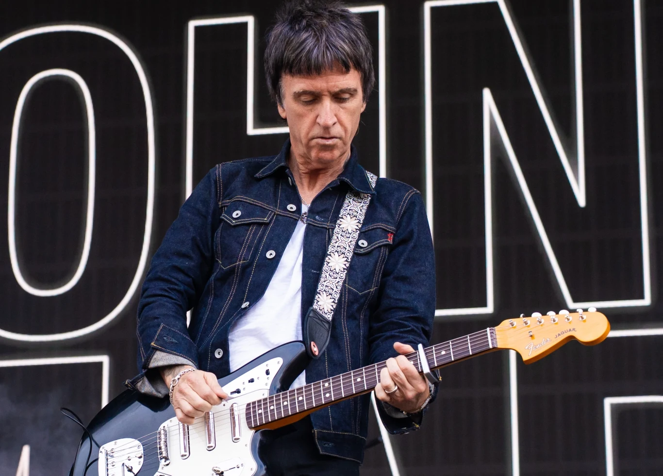 Johnny Marr apeluje do firmy kurierskiej o odzyskanie zamówionych gitar Mężczyzna grający na białej gitarze elektrycznej, ubrany w ciemną kurtkę jeansową, skupiony na grze, w tle duży napis na czarnym tle.
