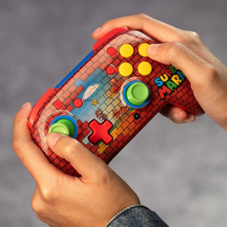 Kolorowy kontroler do gier inspirowany motywem Super Mario, trzymany w obu dłoniach, ozdobiony charakterystycznym wzorem pikselowej cegły oraz napisami, z widocznymi kolorowymi przyciskami i joystickami.