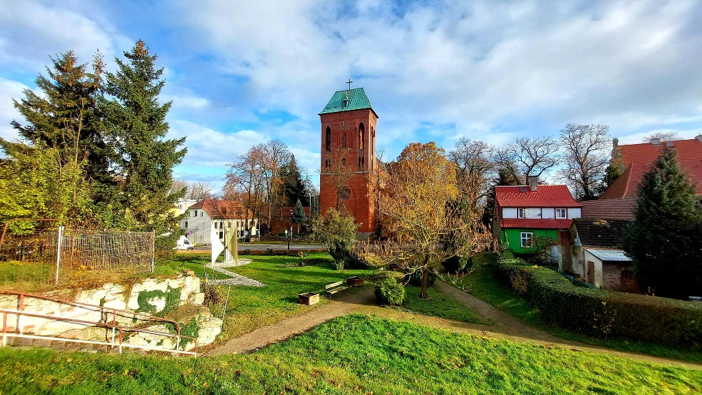 Kamień Pomorski Wieża kościoła z cegły otoczona zielenią, w tle tradycyjne domy i drzewa, wyraźne alejki oraz przyjemnie utrzymany teren zielony pod błękitnym niebem z chmurami.