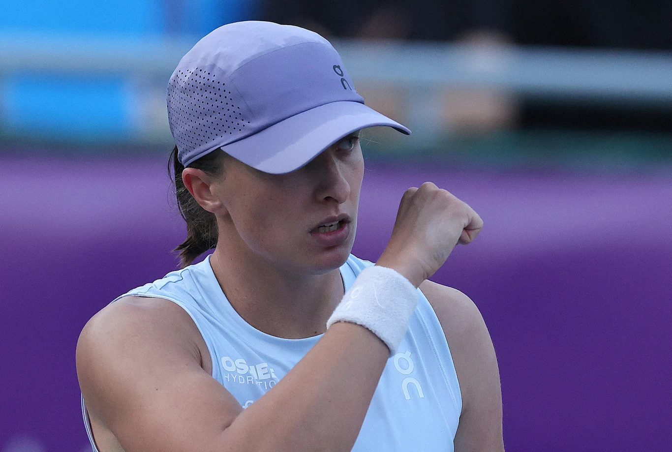 Iga Świątek Tenisistka w jasnej sportowej koszulce i fioletowej czapce wykonuje gest zaciśniętej pięści w trakcie meczu, z wyraźną determinacją na twarzy, w tle rozmyte fioletowe i niebieskie barwy kortu.
