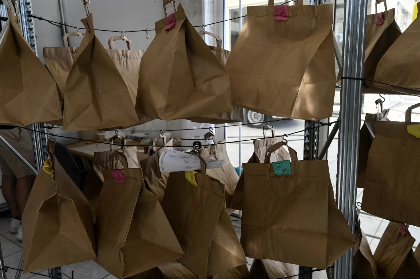 Papierowe torby zawieszone na metalowym regale, każda torba opatrzona kolorowym kawałkiem papieru lub etykietą. Torby są zamknięte, uporządkowane, ustawione w kilku rzędach.