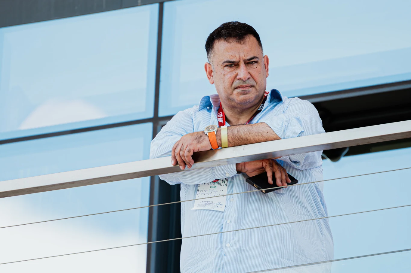 Alex Haditaghi Mężczyzna w średnim wieku w błękitnej koszuli stoi na balkonie i opiera się o balustradę, trzymając w jednej ręce telefon komórkowy; w tle szklana fasada nowoczesnego budynku.