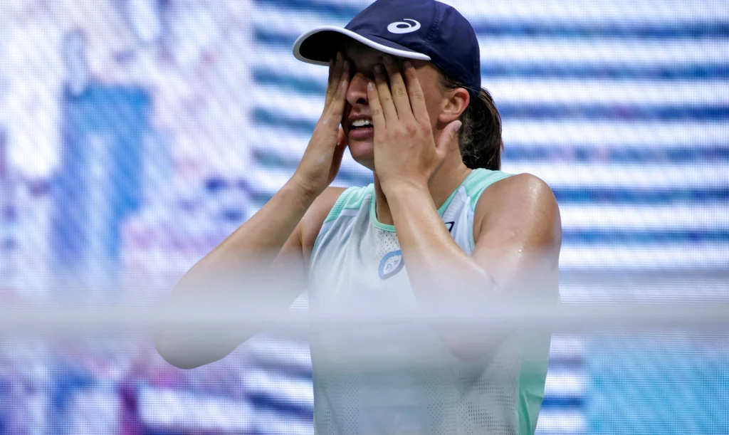 Iga Świątek Sportowiec ubrany w sportowy top i czapkę z daszkiem zasłania twarz dłońmi, wyrażając silne emocje tuż po zakończeniu meczu tenisowego