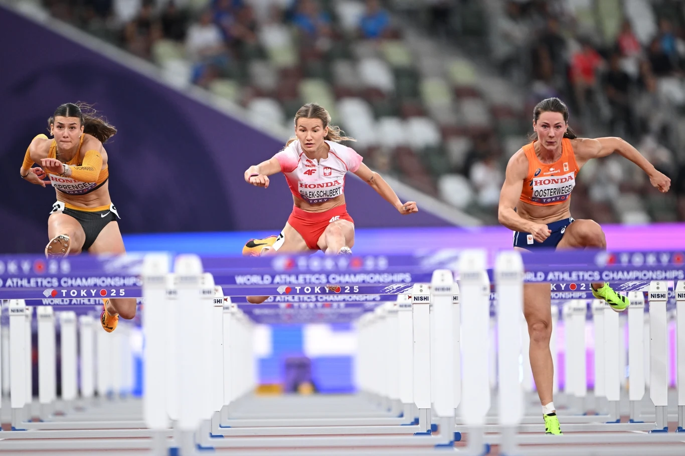Adrianna Sułek-Schubert Trzy lekkoatletki pokonujące płotki podczas zawodów lekkoatletycznych na stadionie, z dynamicznym ujęciem skoku nad przeszkodami, widoczne stroje klubowe oraz napisy na torach i stadionie.