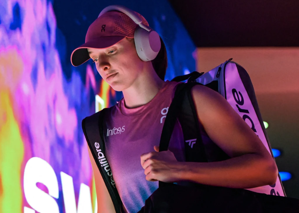 Młoda kobieta ubrana w sportowy strój i czapkę z daszkiem, z dużymi słuchawkami na uszach oraz torbą sportową na ramieniu, idzie obok kolorowej, świetlistej ściany.