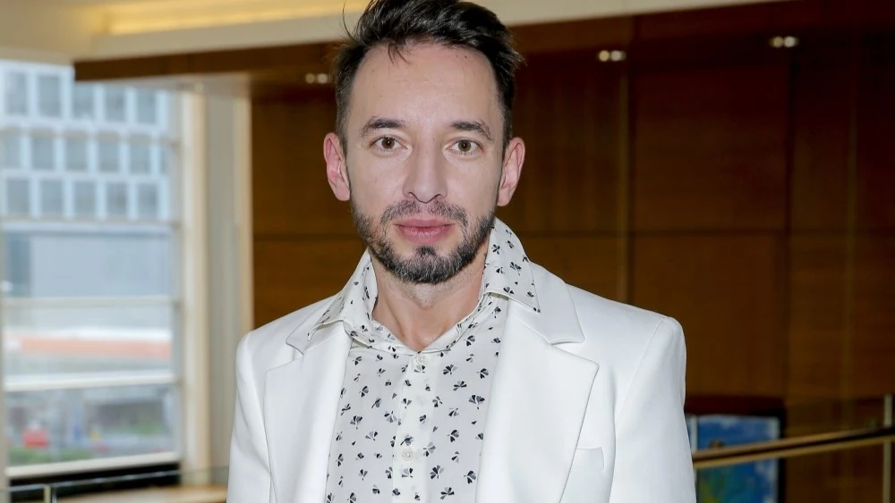 Adam Konkol Mężczyzna z zarostem w białej marynarce i koszuli w drobny wzór, stojący we wnętrzu nowoczesnego budynku z dużymi oknami.