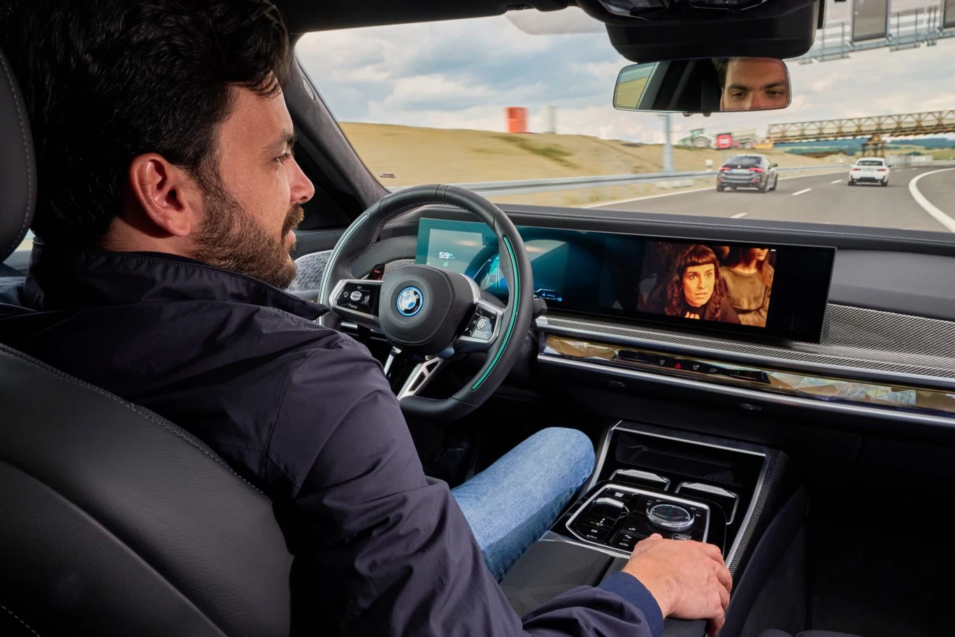 Klienci nie chcą płacić abonamentu za dodatkowe usługi w autach Mężczyzna siedzi za kierownicą nowoczesnego samochodu BMW podczas jazdy autostradą, na dużym ekranie przed kierowcą wyświetlany jest film, a droga za szybą prowadzi przez łagodny zakręt.