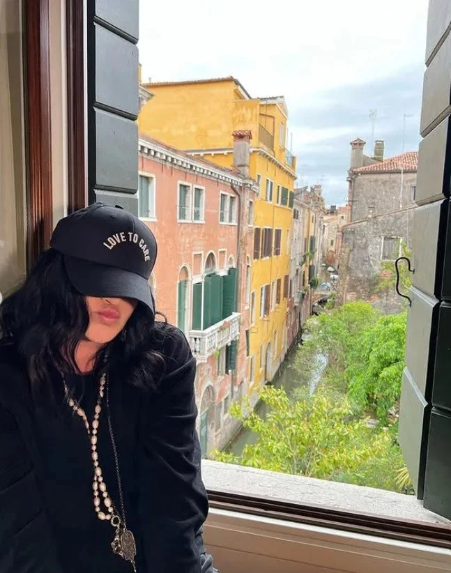 Kayah w swoim domu w Wenecji Młoda kobieta ubrana na czarno z długimi ciemnymi włosami, siedzi na parapecie okna, na głowie ma czapkę z napisem „LOVE TO SAY”, za nią widok na wenecki kanał oraz kolorowe kamienice