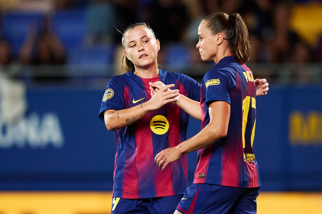 Dwie piłkarki w granatowo-bordowych strojach drużyny FC Barcelona wykonujące gest sportowego uścisku dłoni na boisku w trakcie meczu, w tle zamazana publiczność i fragment trybun.