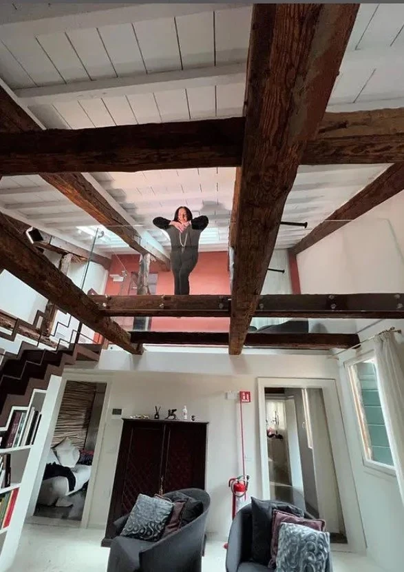 Dom Kayah w Wenecji Nowoczesne wnętrze mieszkania z antresolą, przeszkloną podłogą oraz eksponowanymi drewnianymi belkami stropowymi, na której stoi kobieta w ciemnym stroju. W dolnej części kadru widoczne dwa fotele, schody na antresolę oraz jasne ściany.