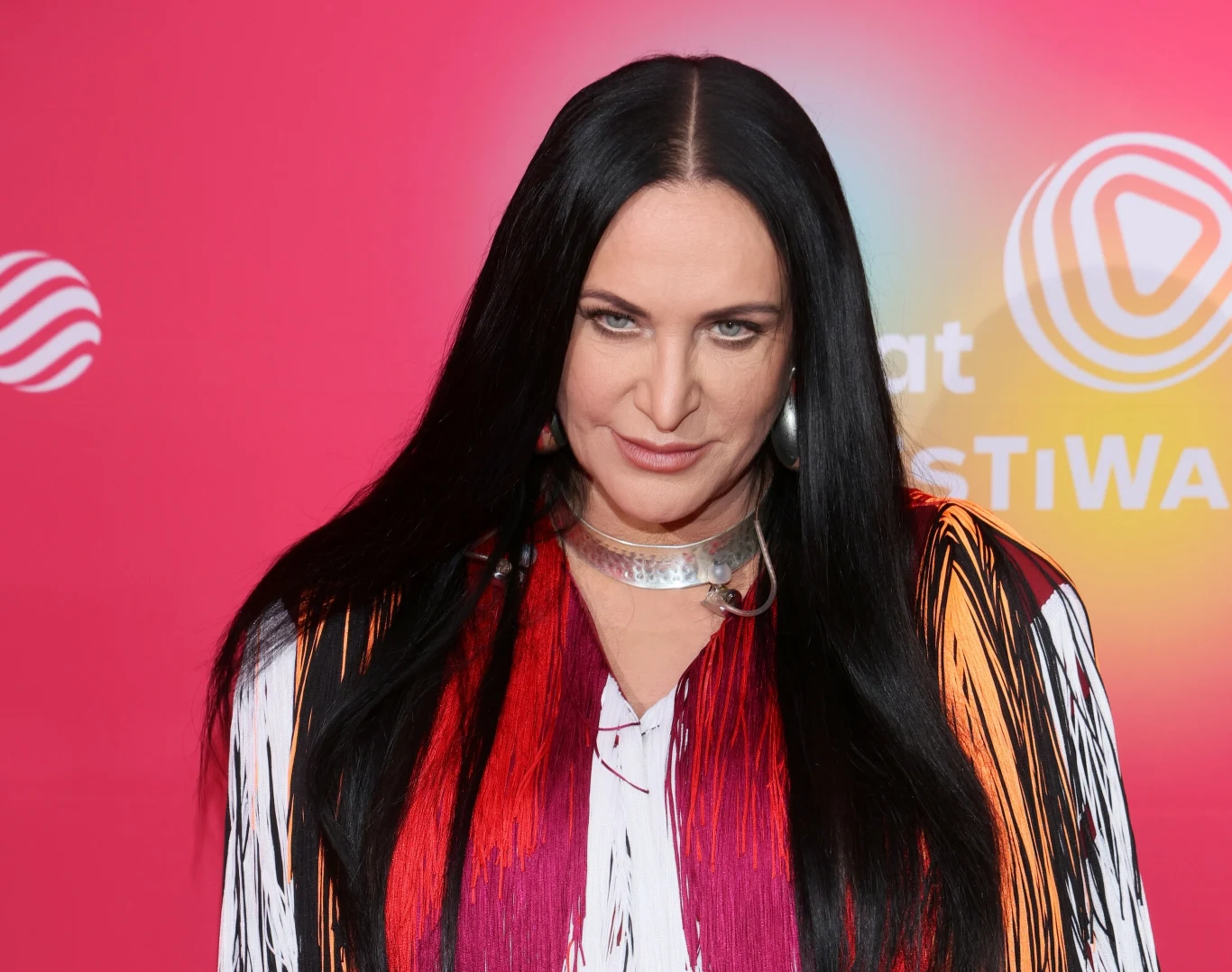 Kayah Kobieta o długich, czarnych włosach ubrana w kolorową, wzorzystą bluzkę, stoi na tle różowej ścianki z logotypami.