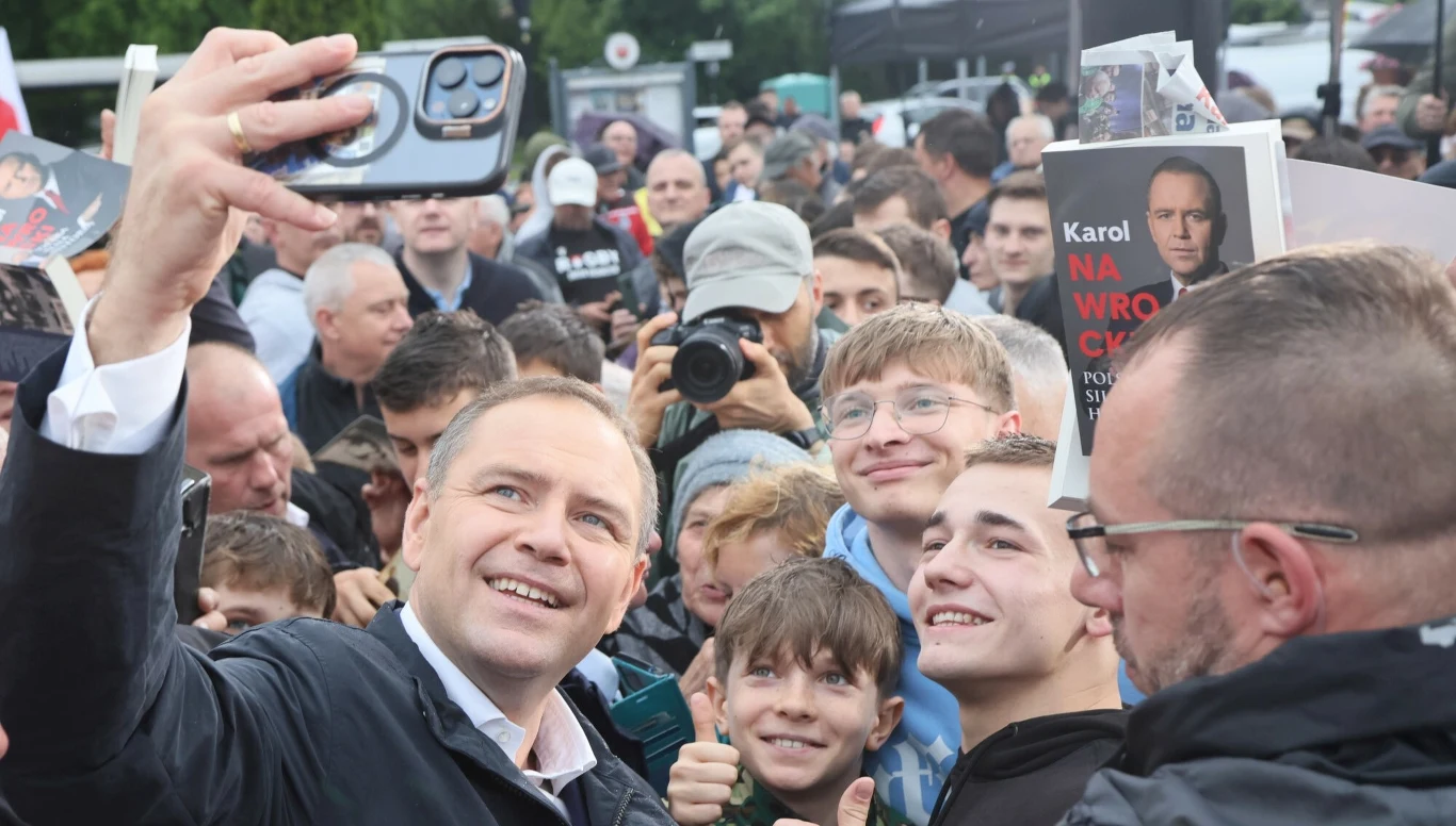 Prezydent RP Karol Nawrocki Tłum ludzi zgromadzony wokół mężczyzny robiącego selfie z grupą uśmiechniętych osób, w tle widoczne transparenty z książką i twarzami uczestników, wokół obecni fotoreporterzy i media.