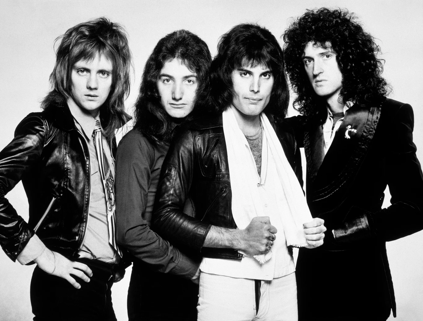 Queen w połowie lat 70. - od lewej: Roger Taylor, John Deacon, Freddie Mercury i Brian May Czterech mężczyzn o długich włosach stoi blisko siebie na tle jasnego tła, wszyscy ubrani są w ciemne ubrania, a dwaj z nich mają ręce skrzyżowane na piersi.