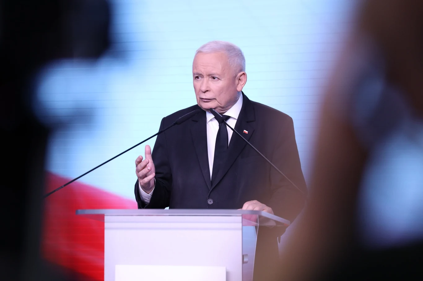 Prezes PiS Jarosław Kaczyński podczas konferencji prasowej Starszy mężczyzna w garniturze przemawia przy przezroczystej mównicy, w tle rozmyty jasny ekran z elementami czerwieni i bieli, dwie mikrofony skierowane w jego stronę.