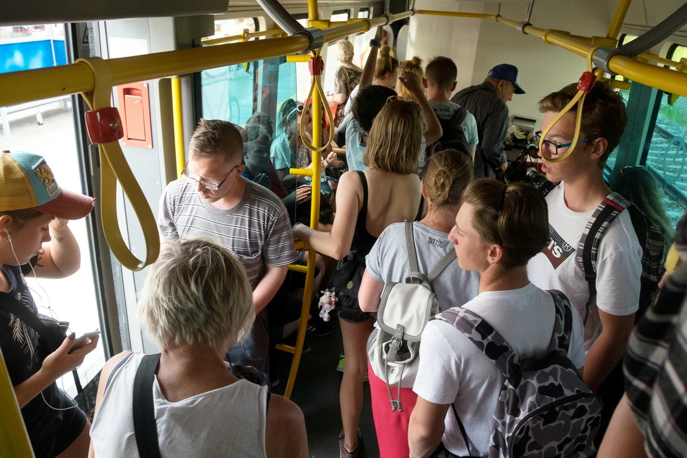 Tłok w autobusie, pasażerowie o różnym wieku stojący blisko siebie, część osób z plecakami, kilka osób korzysta z telefonu, przez otwarte drzwi wpada światło dzienne.