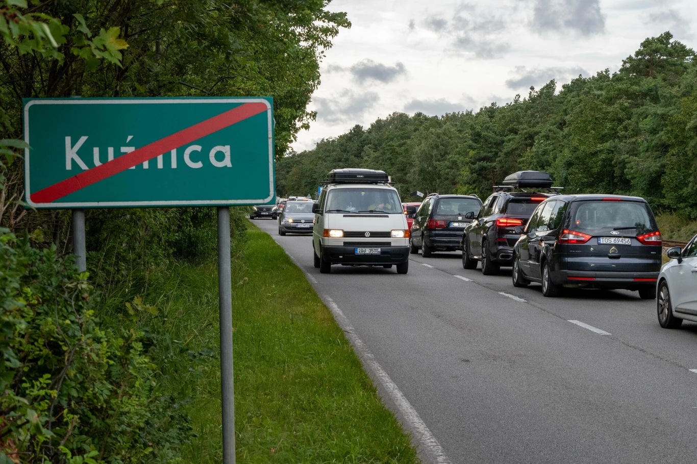Korek samochodowy na dwupasmowej drodze otoczonej zielenią i lasem, po lewej stronie znak drogowy z przekreśloną nazwą miejscowości Kuźnica