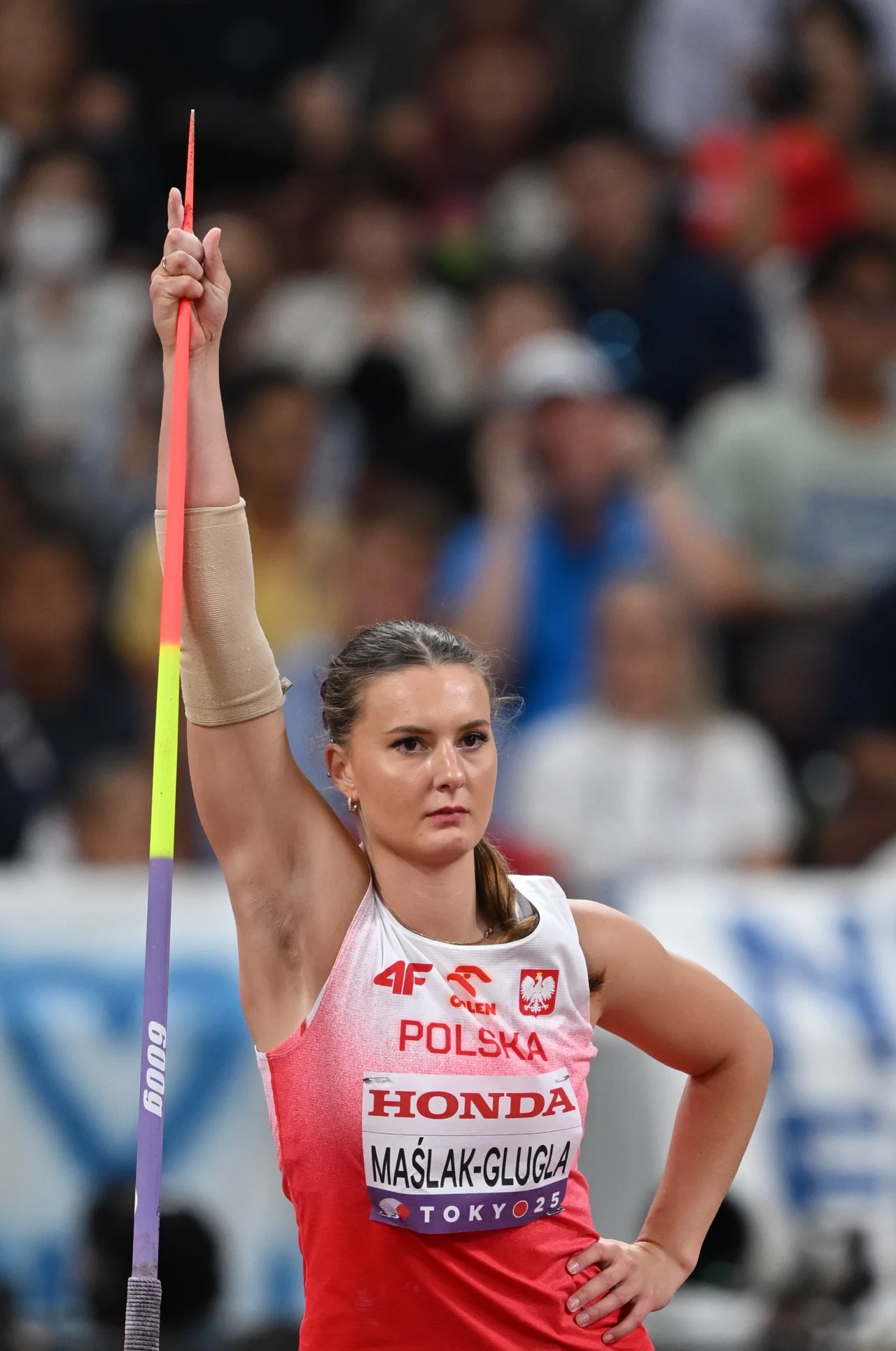 Zawodniczka reprezentująca Polskę w lekkoatletyce stoi skoncentrowana na stadionie, trzymając dzidę w pozycji pionowej przed rzutem, ubrana w sportowy strój z napisem Polska i nazwiskiem na koszulce, w tle rozmyta publiczność.
