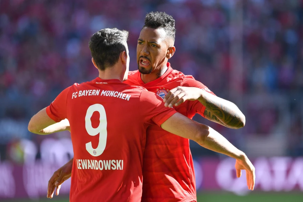 Jerome Boateng i Robert Lewandowski z czasów wspólnej gry w Bayernie Monachium Piłkarze Bayernu Monachium w czerwonych strojach świętują zdobycie gola na tle stadionu wypełnionego publicznością, jeden zawodnik przytula drugiego z widocznym entuzjazmem.