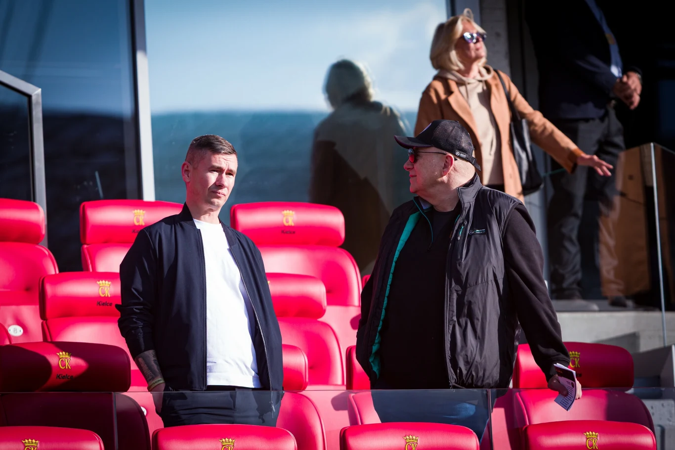 Dwóch mężczyzn stoi pośród czerwonych foteli stadionowych, rozmawiając ze sobą; w tle widoczna kobieta w okularach przeciwsłonecznych.