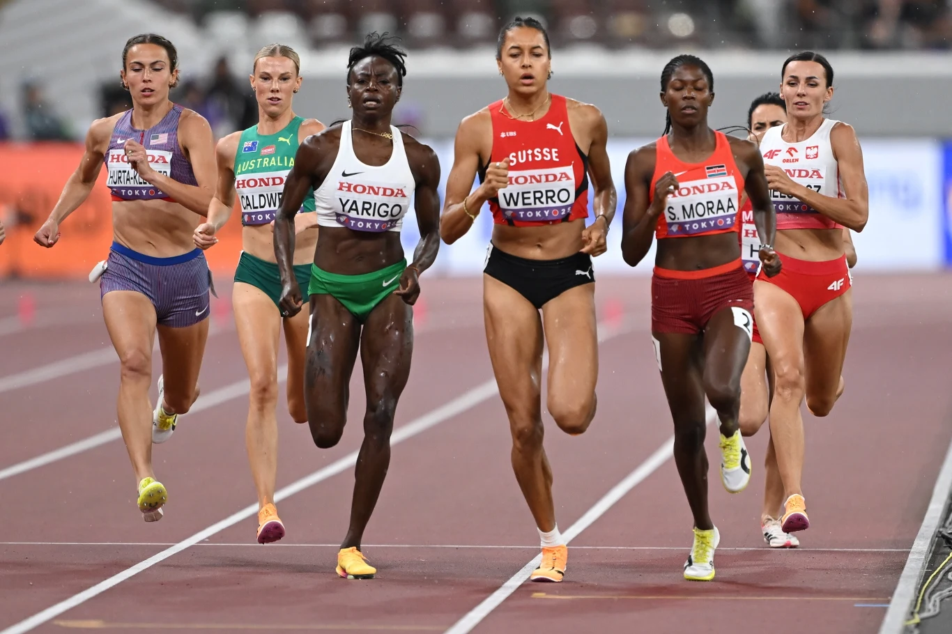 Grupa zawodniczek biegnie po bieżni lekkoatletycznej podczas wyścigu, wszystkie są w sportowych strojach, skoncentrowane i rywalizują o pierwsze miejsce, w tle widać trybuny stadionu